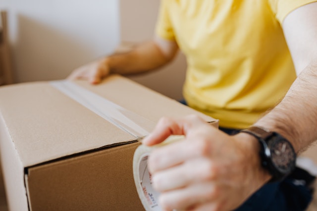 someone-sealing-a-carton-box-while-stil-holding-the-tape-wearing-a-yellow-shirt-and-a-black-watch